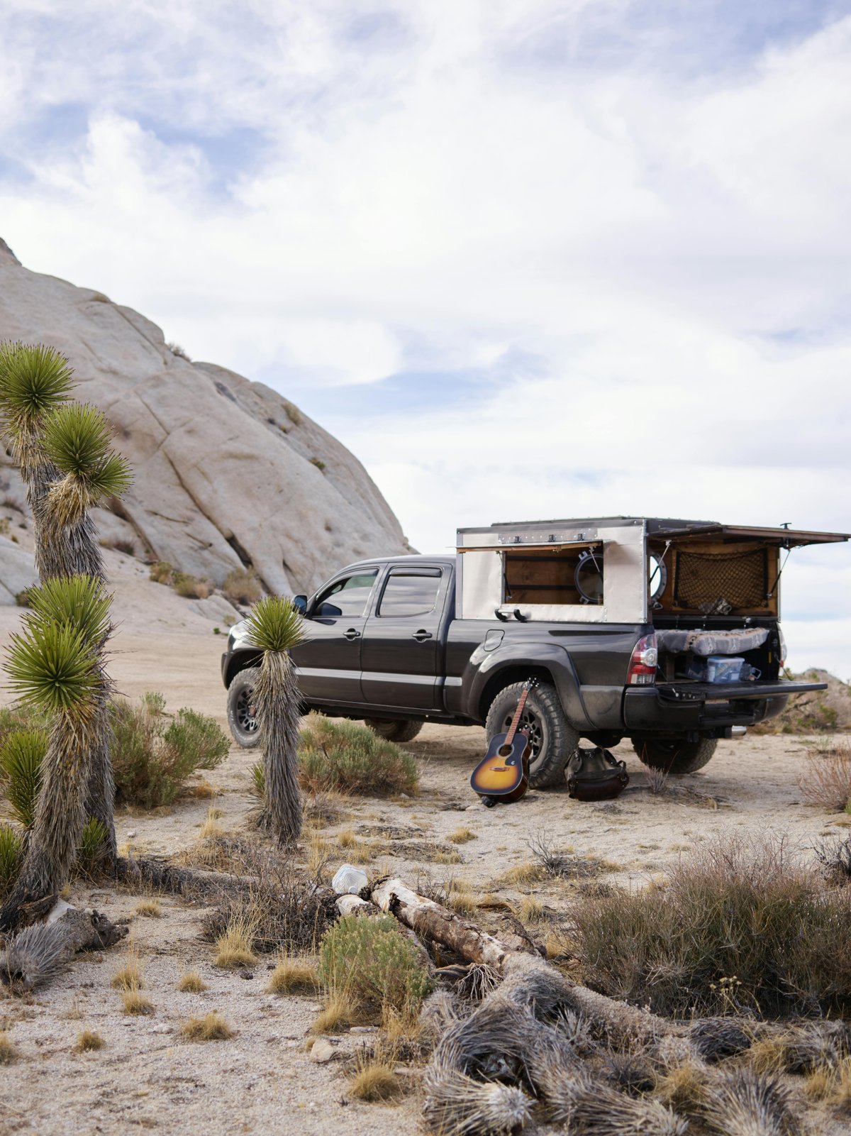Pickup truck parked in desert with guitar and open camper, surrounded by Joshua trees.